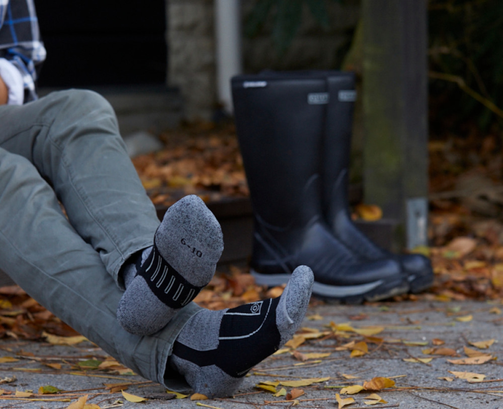 A person wearing grey and black socks sits on an outdoor surface covered with fallen leaves. Their boots are placed on the ground nearby. Only the persons legs and feet are visible.
