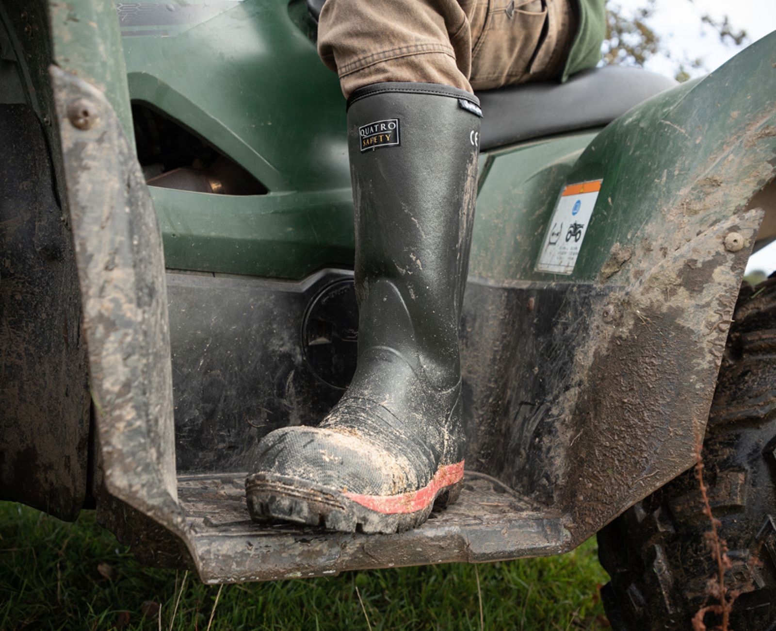 A person wearing muddy green rubber boots rests their foot on the step of a dirty green ATV, with grass and mud visible on the ground.