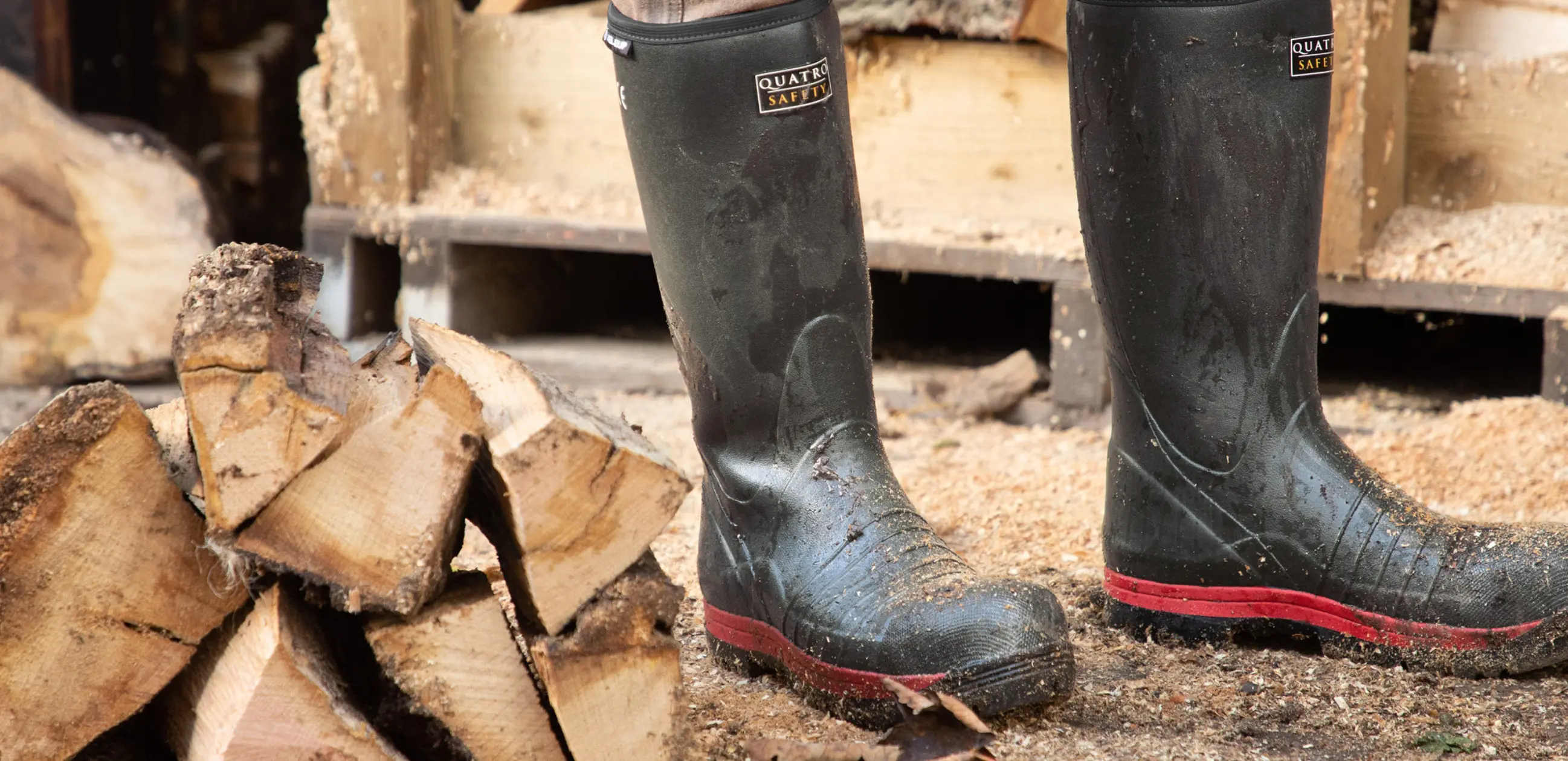A pair of muddy black rubber boots stands on the ground near chopped firewood, with an axe and stacked logs in the background.