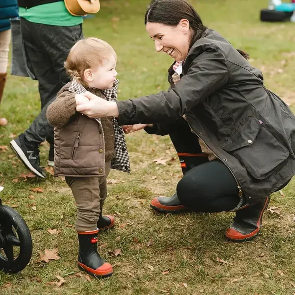 A woman crouches on grass, smiling and holding the hands of a toddler who is standing. Both wear boots and jackets. Fallen leaves are on the ground, and other people and part of a stroller are visible in the background.