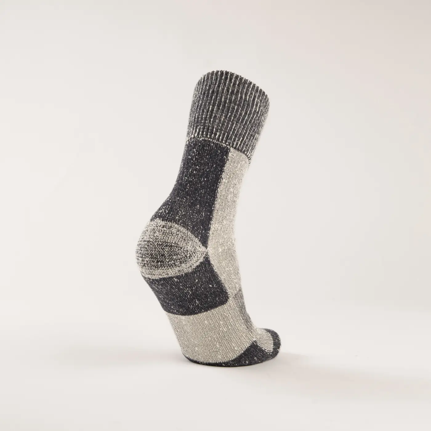 A single grey and white wool sock stands upright on a plain, light background, displaying its heel, toe, and a color-blocked pattern with varying shades of grey and white.