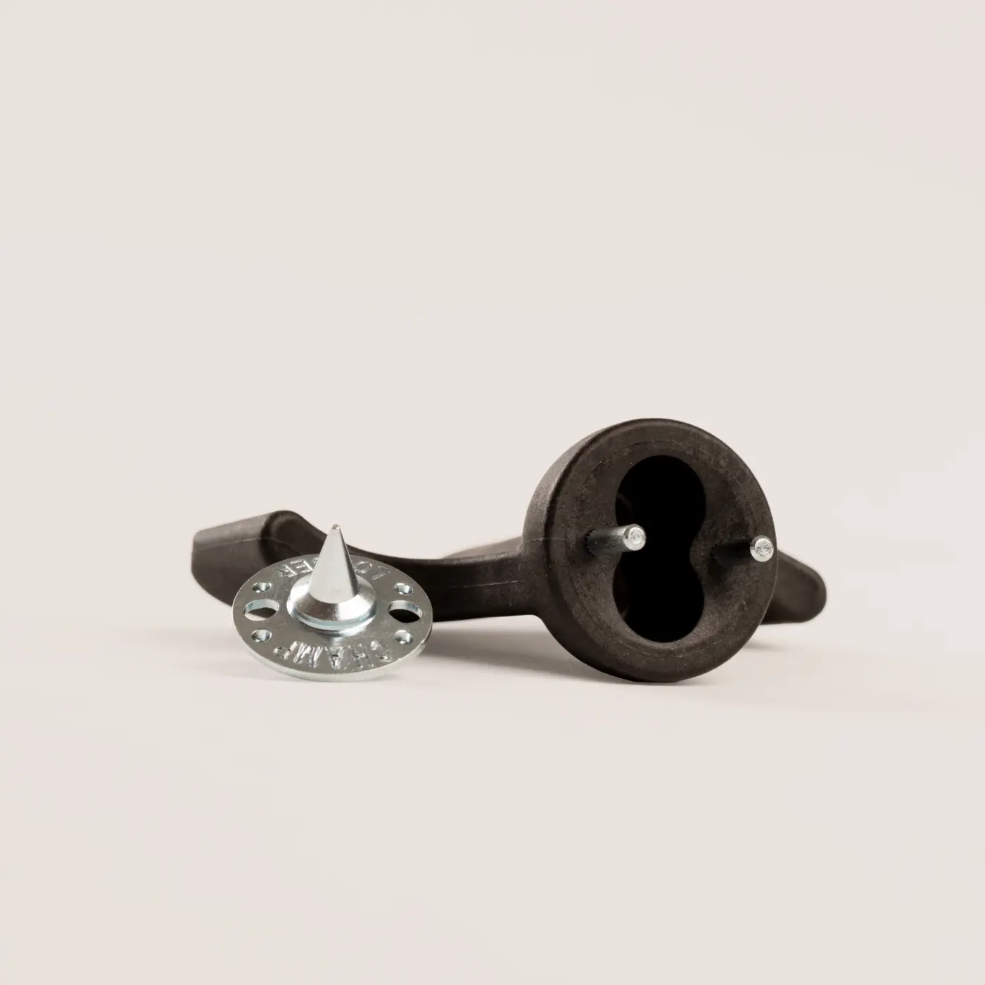 A black plastic tool with a circular base and two prongs lies on its side next to a round silver metal spiked disc with small holes around its edge, both on a plain light surface.