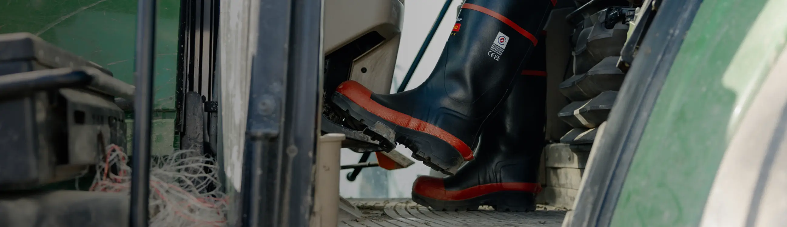 A person wearing black and red safety boots is pressing the pedal inside a green tractor, with netting visible on the floor near the door.