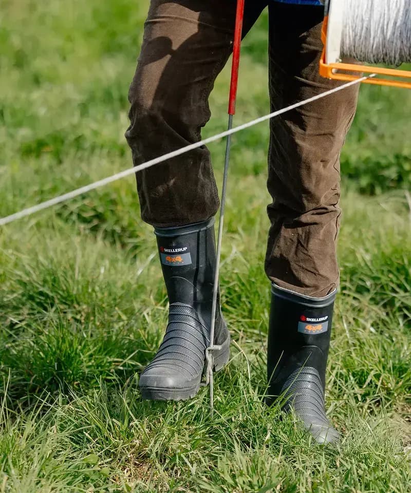 A person wearing black rubber boots and brown pants stands on grass, using a tool to secure a wire fence. Only their legs and boots are visible.