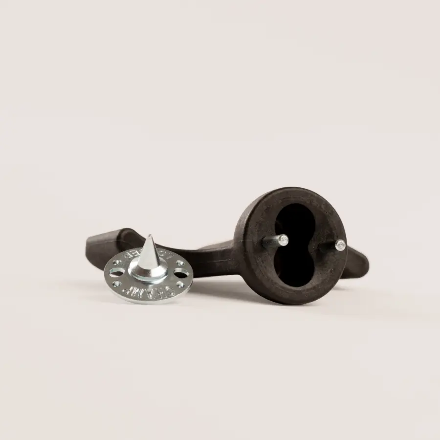 A black plastic tool with a circular base and two prongs lies on its side next to a round silver metal spiked disc with small holes around its edge, both on a plain light surface.