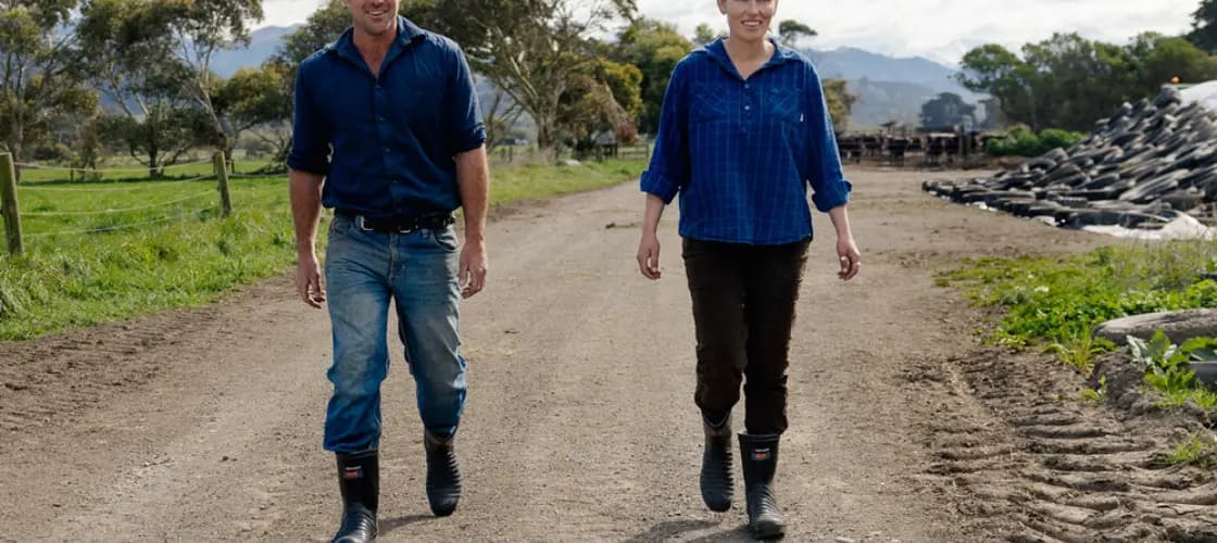A man and a woman wearing boots and casual farm clothes walk side by side on a dirt road in a rural setting with trees and mountains in the background.