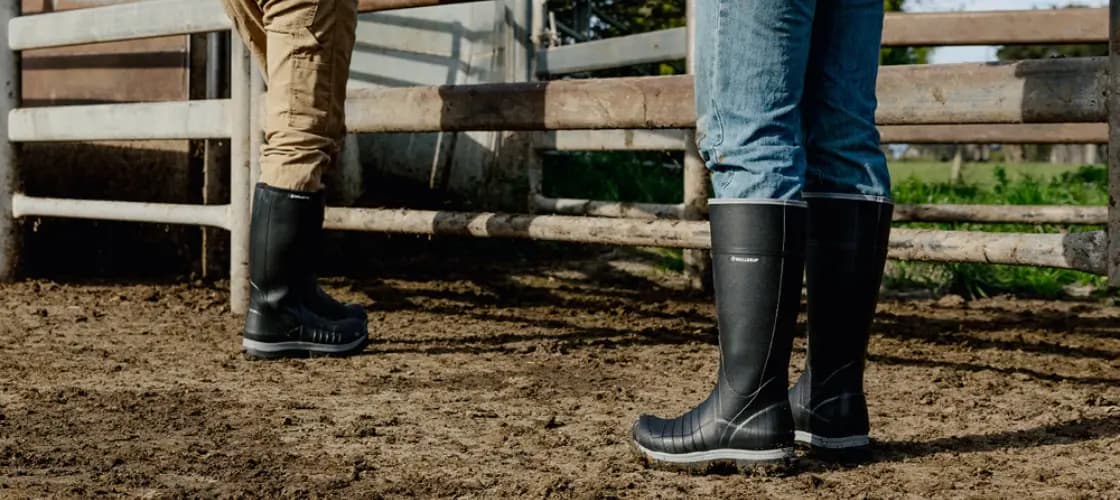 Two people wearing jeans and rubber boots stand on muddy ground near a metal fence, possibly on a farm or ranch. Their upper bodies are mostly out of the frame.