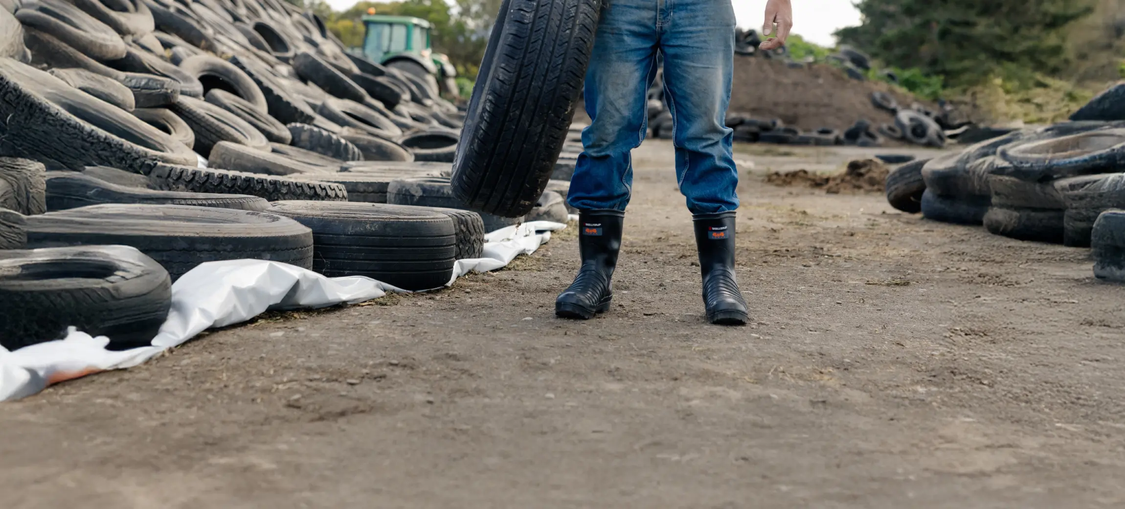 A person in jeans and black boots stands outdoors holding a large tire upright, surrounded by many piled, discarded tires on the ground. Only the lower half of the person is visible.