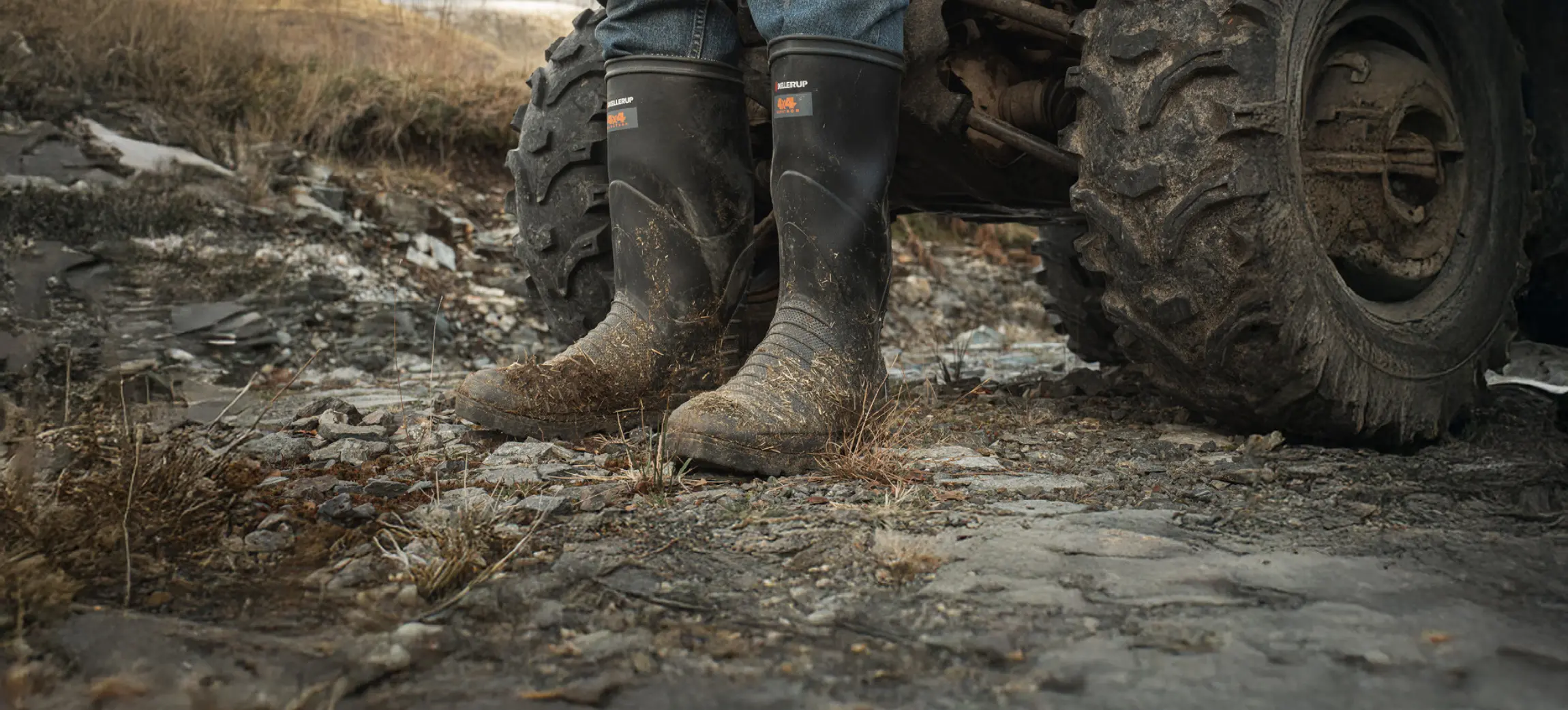 A person wearing muddy black rubber boots and jeans stands next to a large, muddy ATV tire on a rocky outdoor terrain. The scene suggests rural or off-road activity.