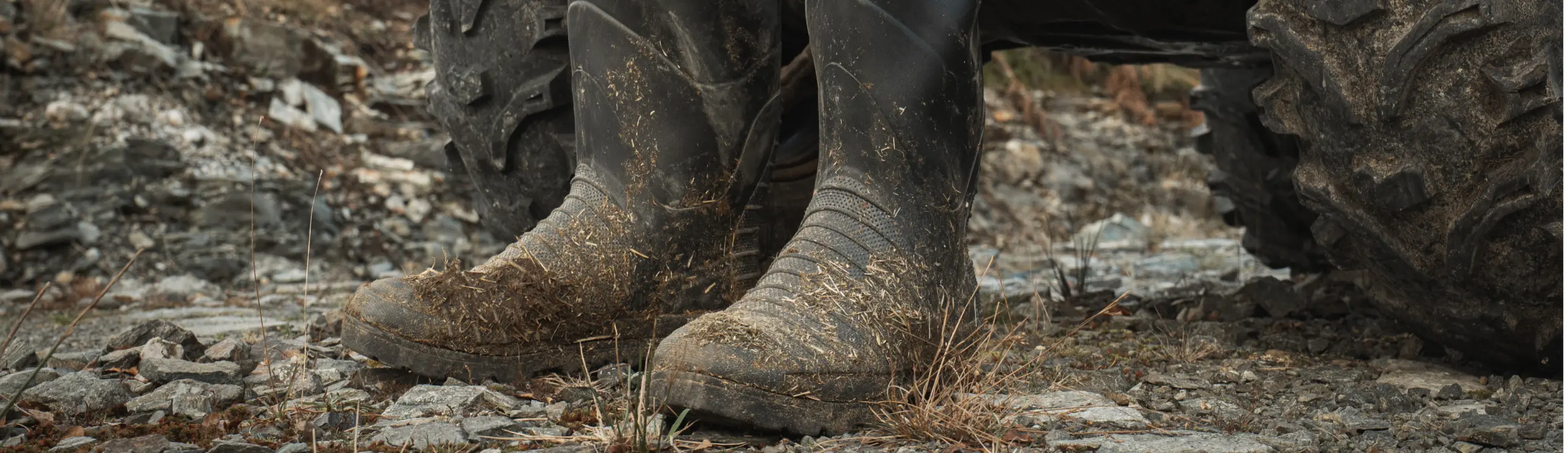 A close-up of a person wearing muddy black rubber boots standing on rocky, uneven ground beside the large tire of an off-road vehicle. Dry grass and stones are visible around their feet.