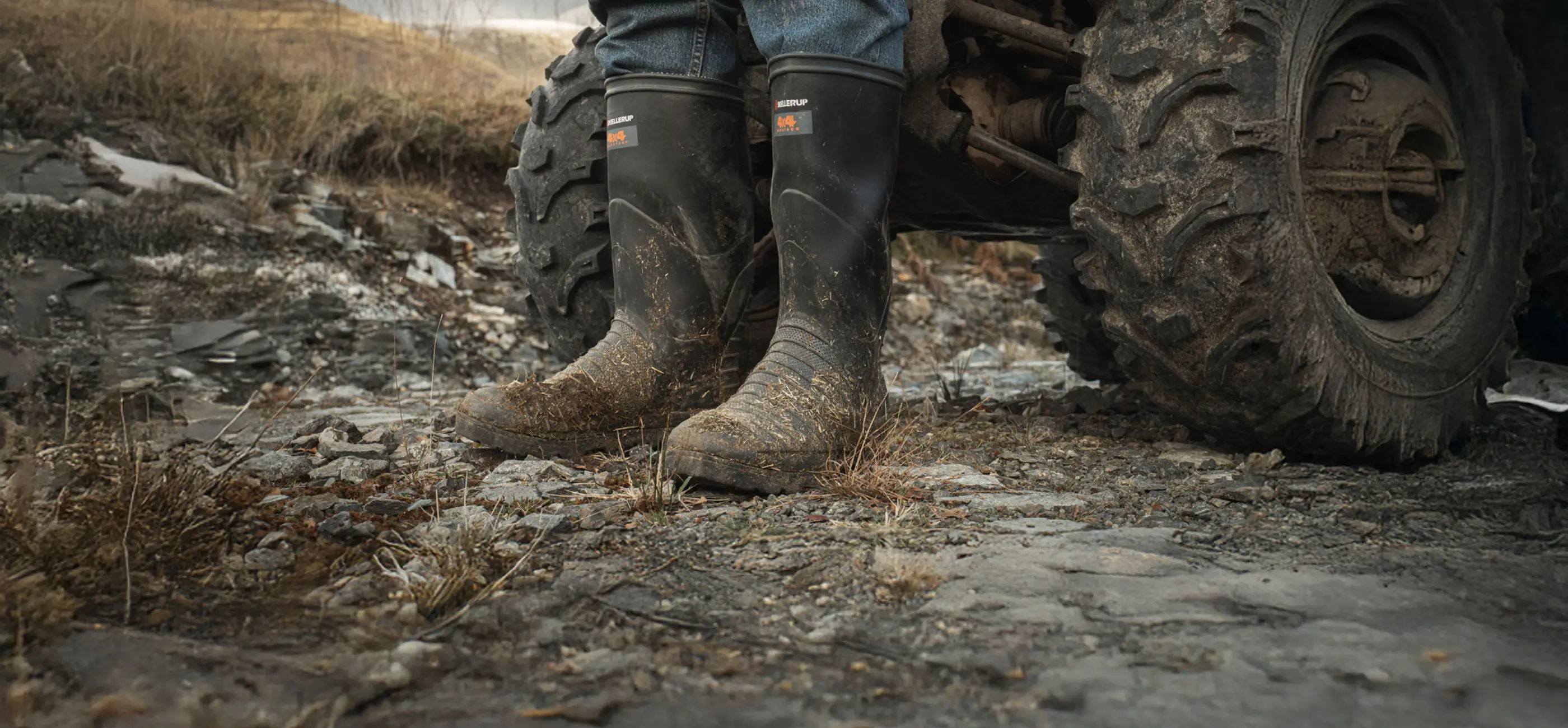 A person wearing muddy black rubber boots and jeans stands next to a large, muddy ATV tire on a rocky outdoor terrain. The scene suggests rural or off-road activity.