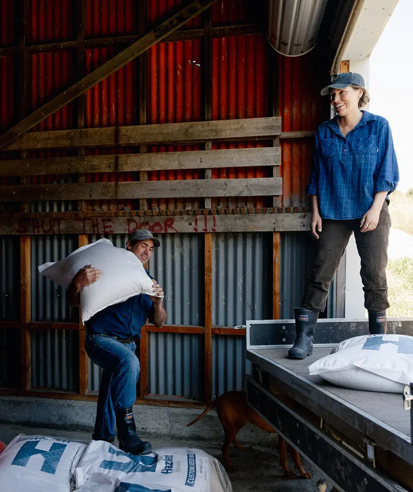 A man carrying a large white sack over his shoulder stands next to stacked bags in a barn, while a woman in a blue shirt and cap smiles at him from a raised platform. A brown dog is partially visible below.