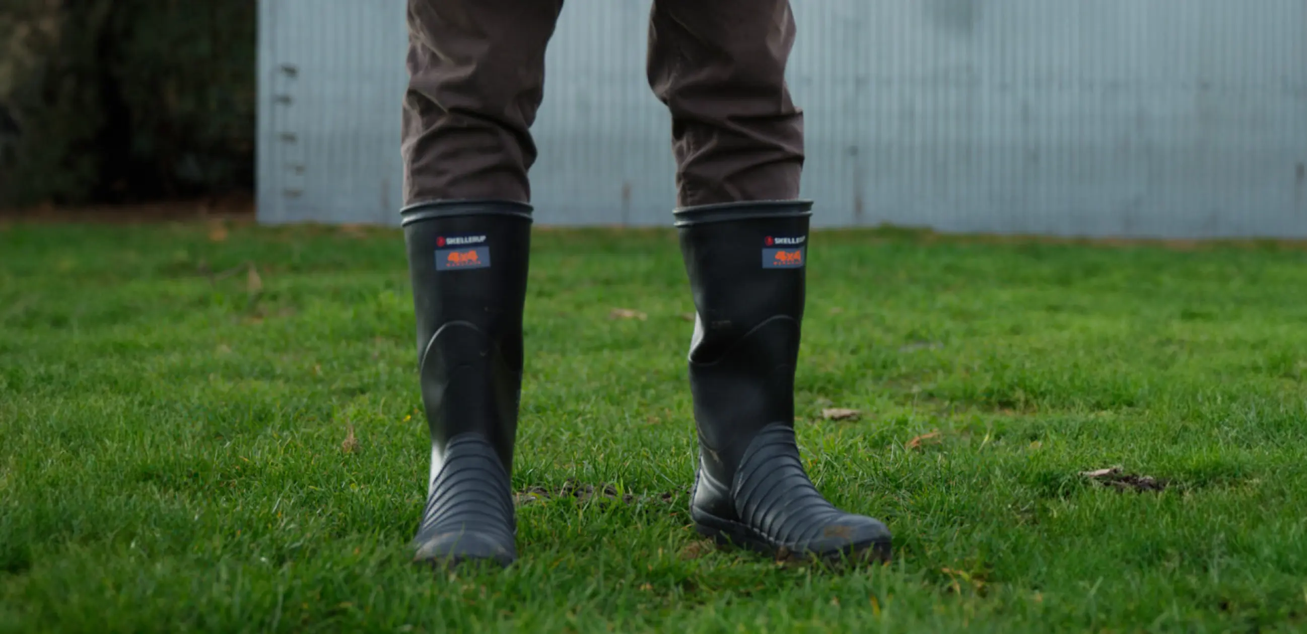 A person standing on green grass wearing black rubber boots and brown pants, with only their legs visible; a corrugated metal wall is in the background.
