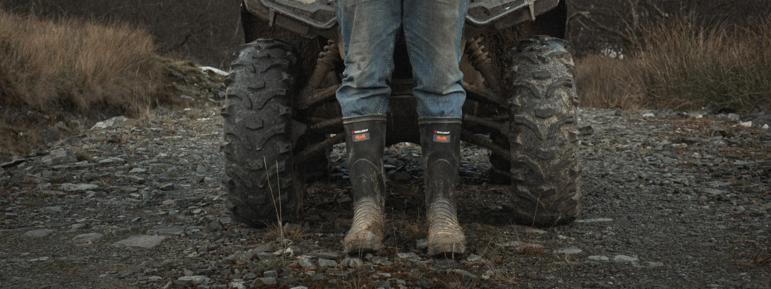 A person wearing muddy boots and rolled-up jeans stands in front of an ATV on a rocky, outdoor trail with dry grass and bushes in the background.