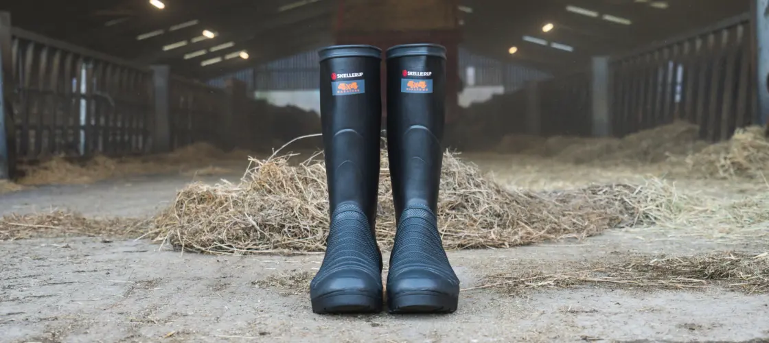 A pair of black rubber boots stand upright on a barn floor, surrounded by hay, with animal pens and overhead lights visible in the background.