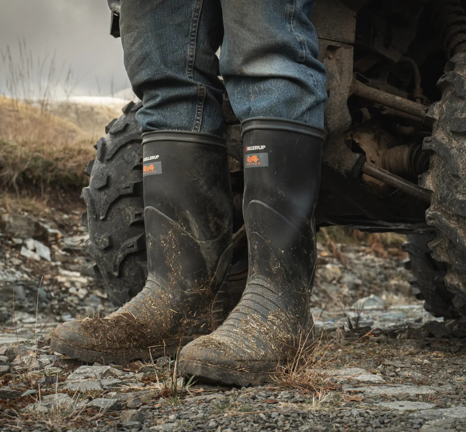 A person wearing muddy black rubber boots and blue jeans stands on rocky ground next to the large tire of an off-road vehicle in an outdoor setting.