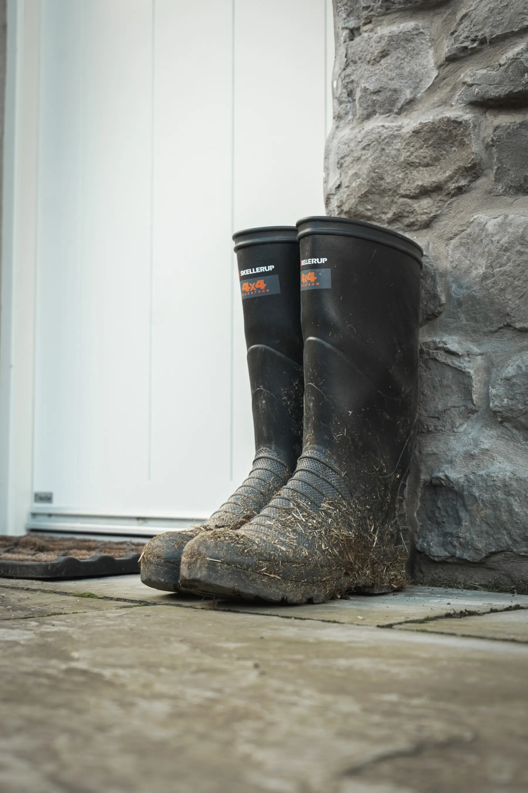 A pair of muddy black rubber boots are standing upright against a stone wall next to a white door on a stone patio.