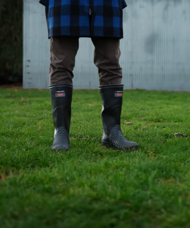 A person wearing brown pants, black rubber boots, and a blue plaid coat stands on green grass in front of a gray metal wall. The photo is cropped at the shoulders.