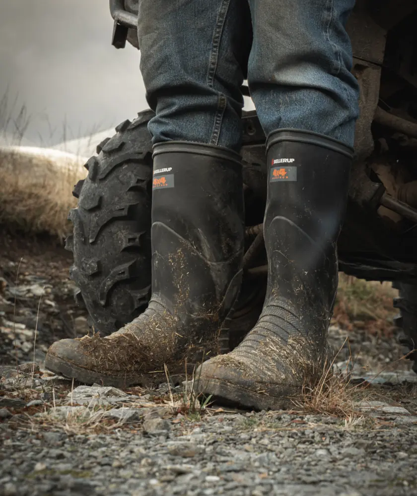A person wearing muddy black rubber boots and blue jeans stands on rocky ground beside an ATV tire on a cloudy day.