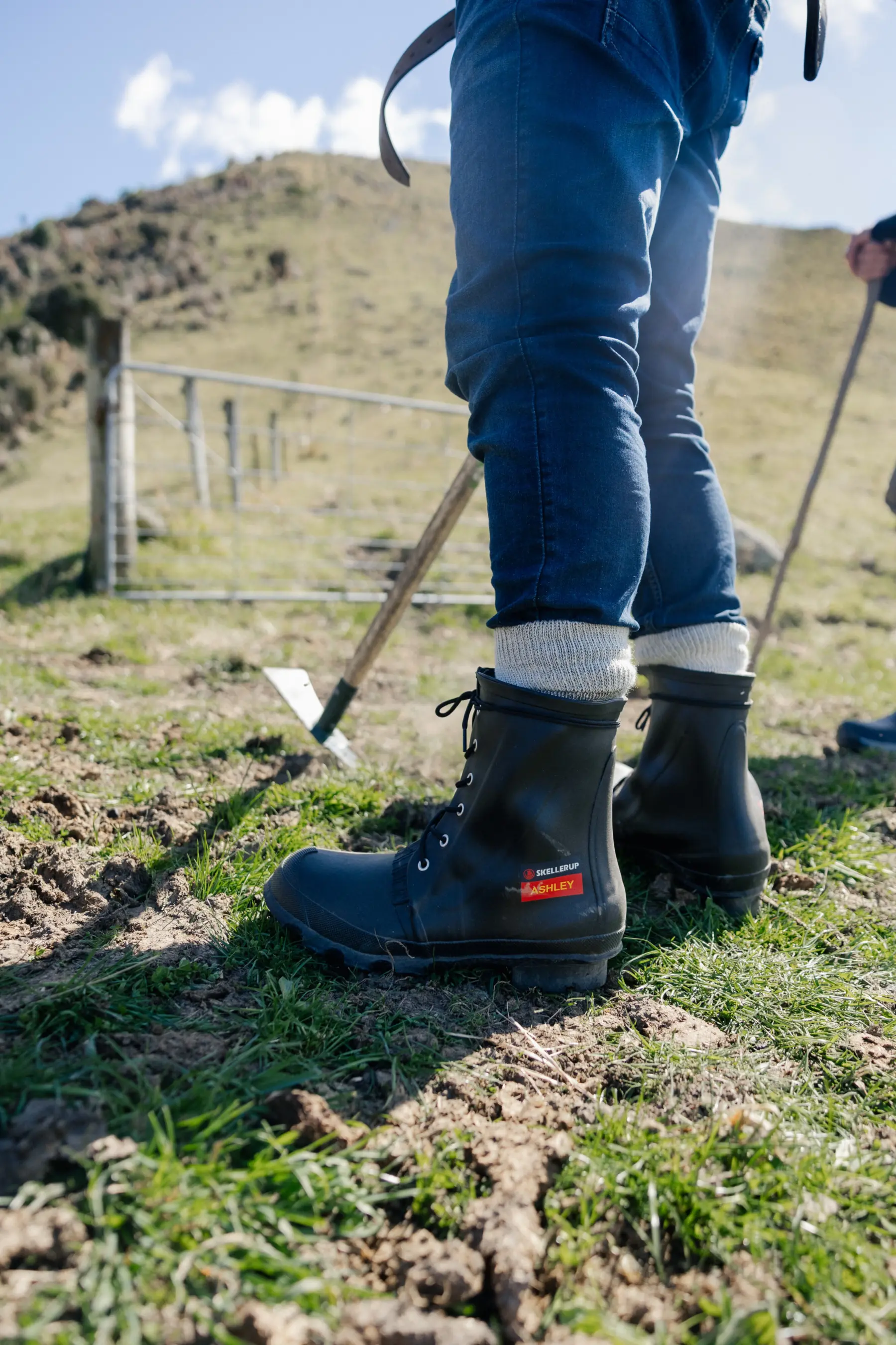 A person in jeans, light socks, and black rubber boots stands on grassy, uneven ground near a metal gate and hilly landscape, holding a wooden stick or walking pole. Another person is partly visible in the background.