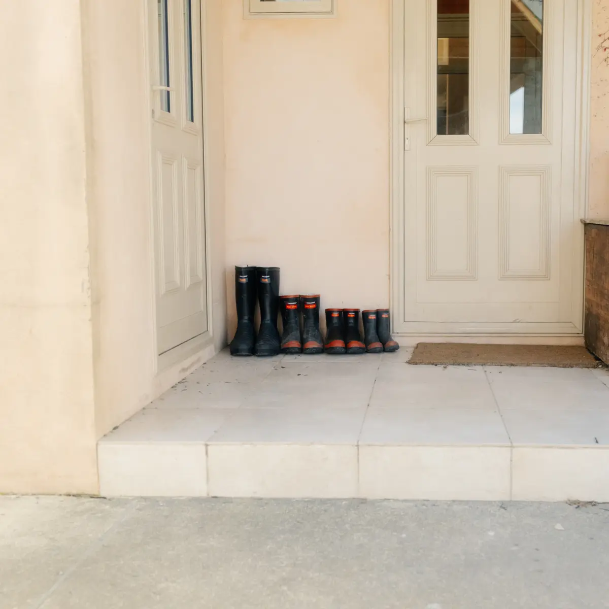 A tiled porch with three pairs of black rubber boots lined up by a door, next to a weathered wooden box and a brown paper bag. The surroundings are beige walls with bare vines.