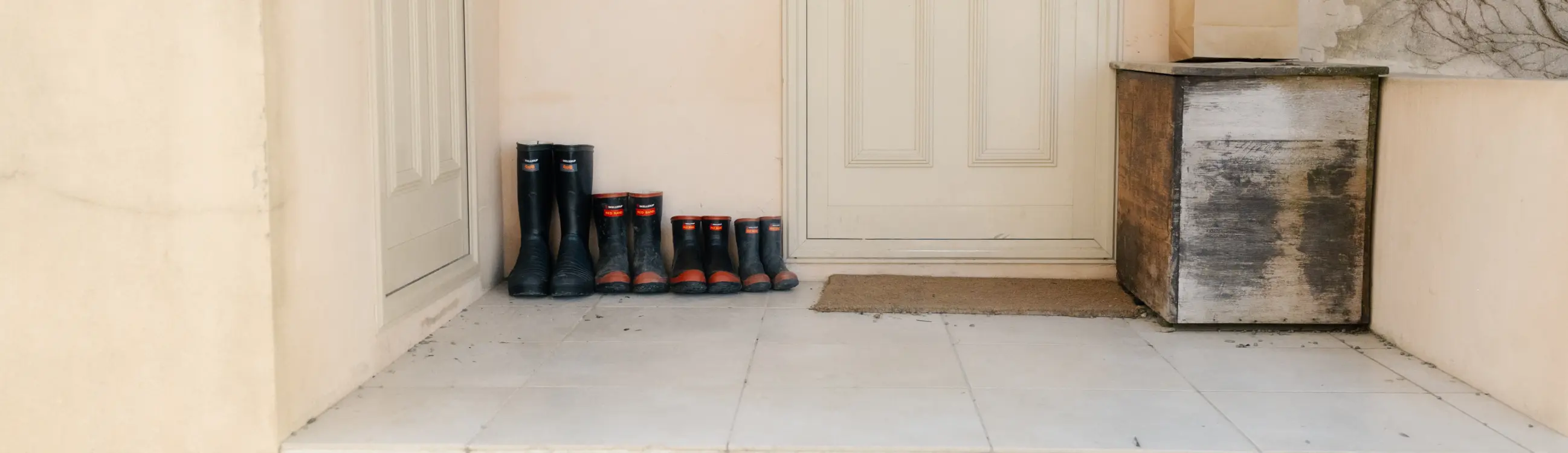 A tiled porch with three pairs of black rubber boots lined up by a door, next to a weathered wooden box and a brown paper bag. The surroundings are beige walls with bare vines.