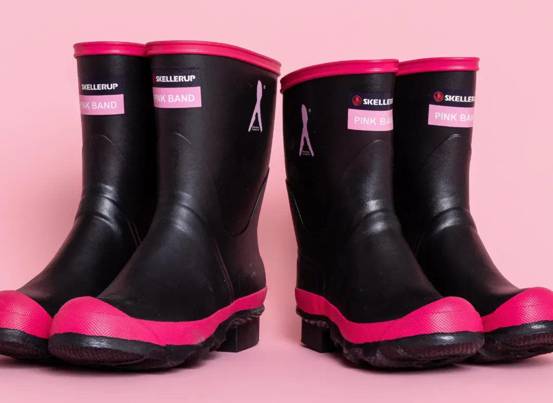Two pairs of black rubber boots with pink soles and trim are placed side by side on a pink background. The boots have labels on the front.
