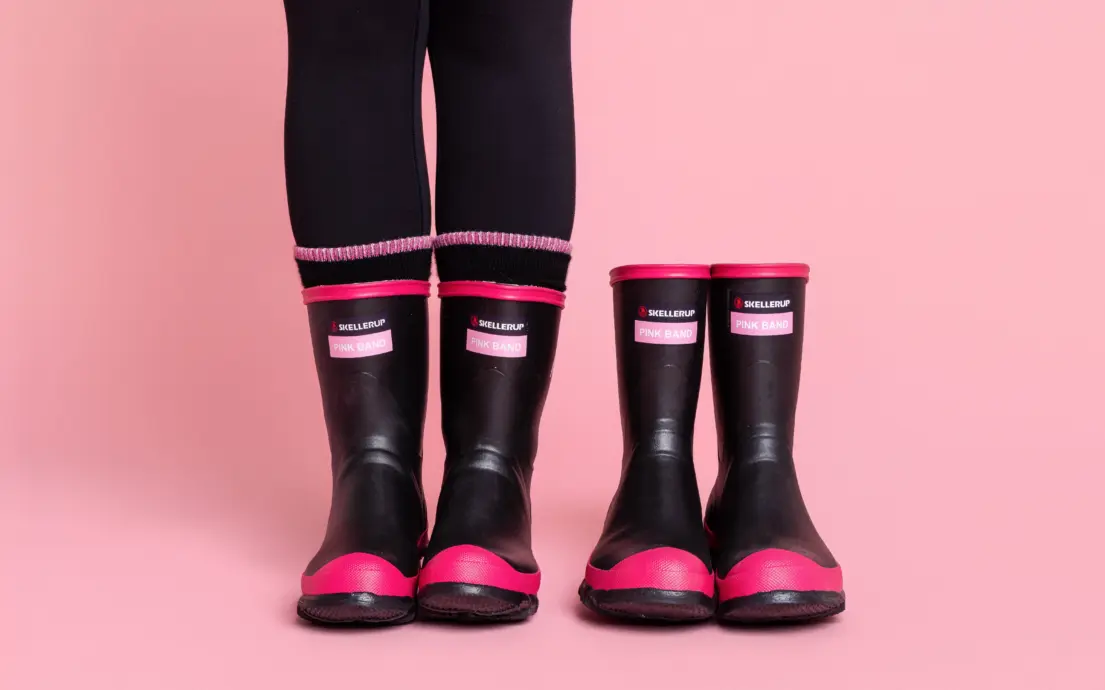Two pairs of black and pink rubber rain boots, one larger and one smaller, are placed side by side on a pink background. A person wearing black leggings and the larger boots stands next to the smaller pair.