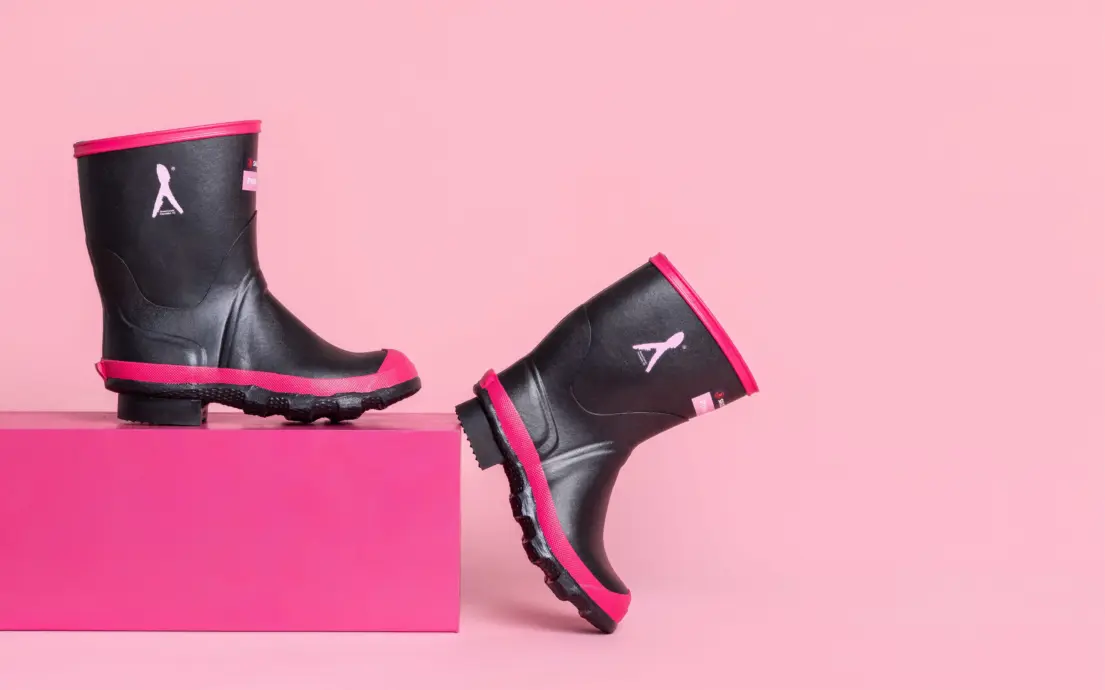 Two black rain boots with pink soles and trim are displayed against a pink background; one boot stands on a pink block while the other is propped up, showing the sole and pink accents.