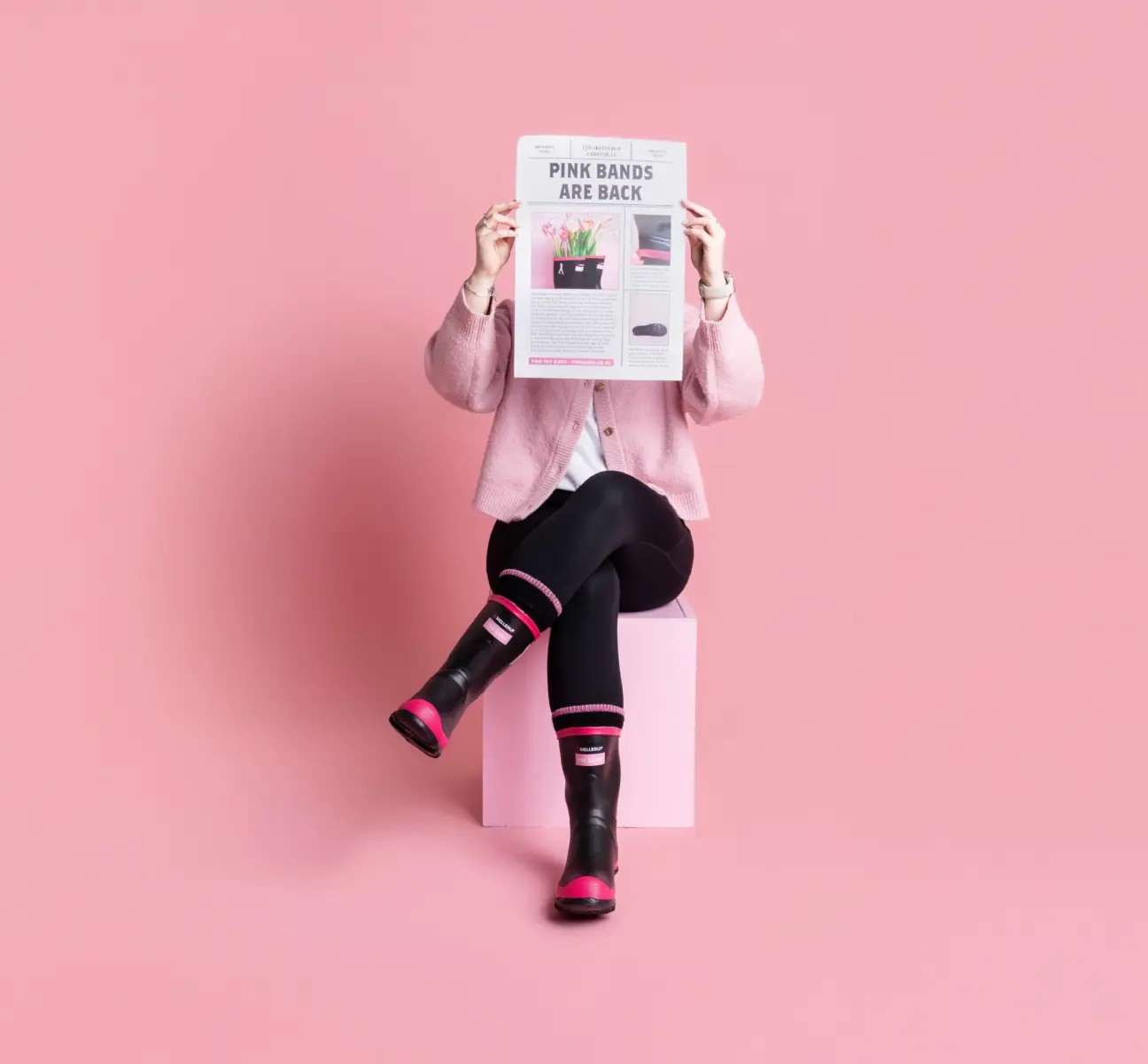 A person wearing black leggings, pink and black boots, and a pink cardigan sits on a pink cube against a pink background, holding up a newspaper that covers their face.