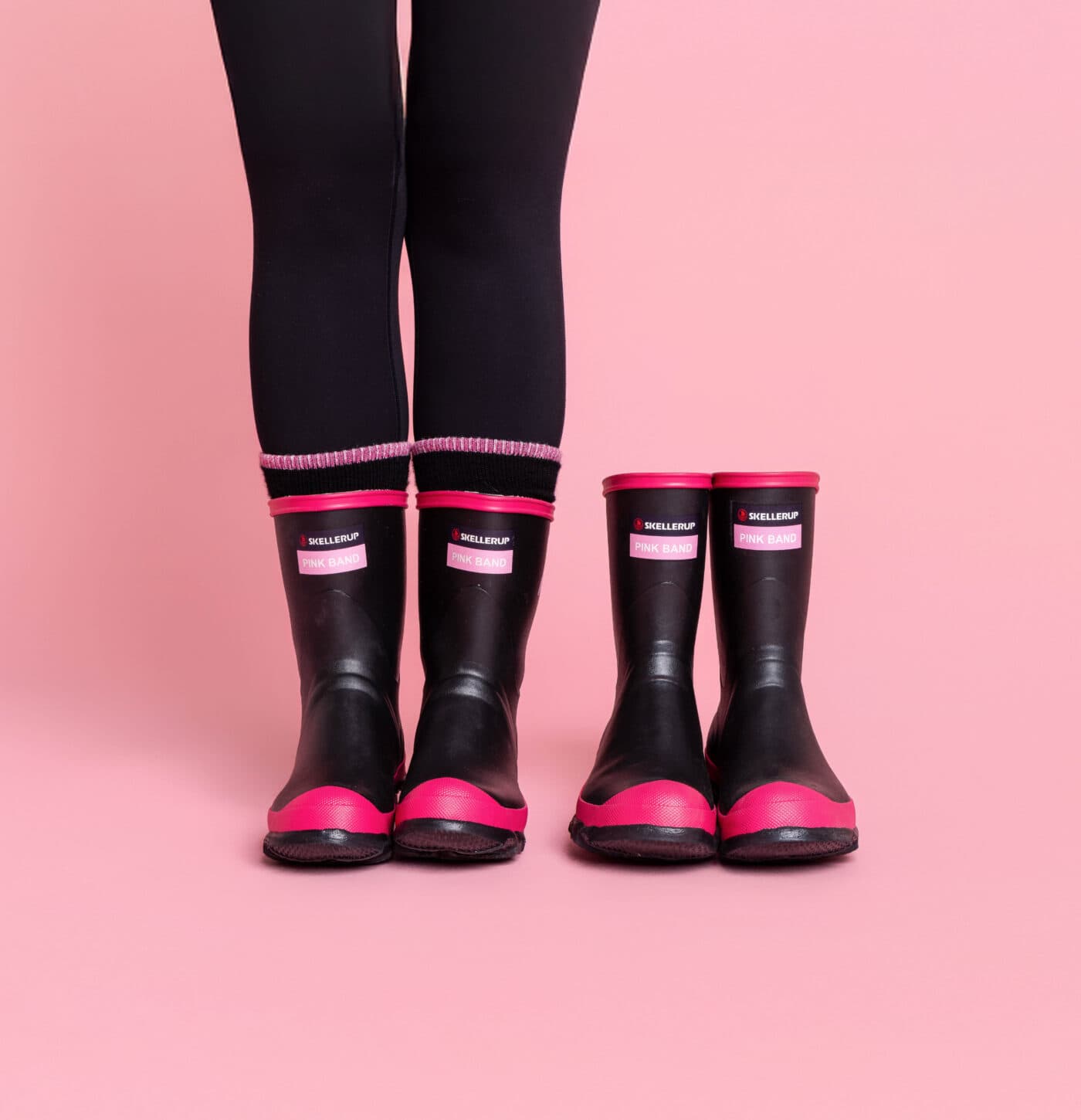 Two pairs of black and pink rubber rain boots, one larger and one smaller, are placed side by side on a pink background. A person wearing black leggings and the larger boots stands next to the smaller pair.