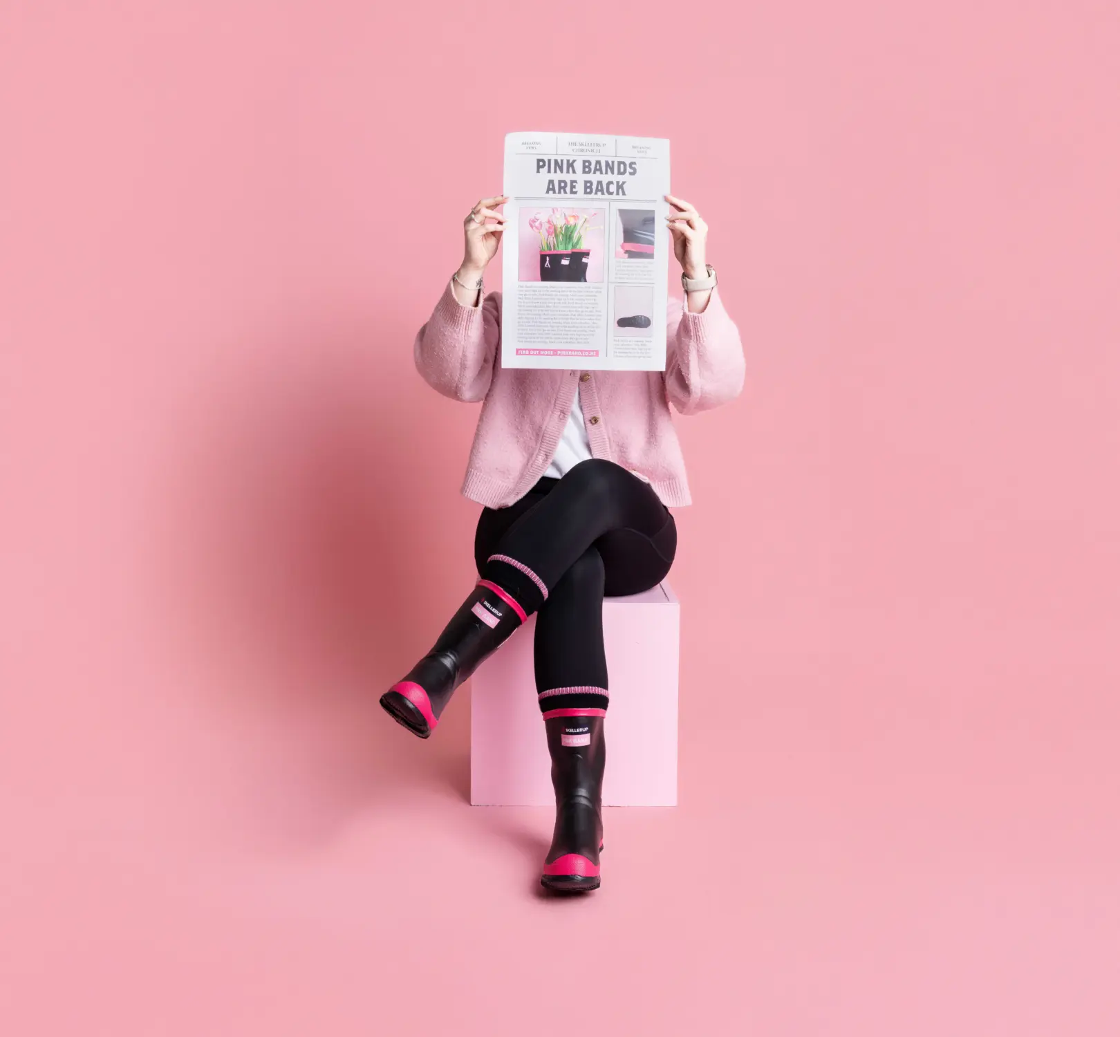 A person wearing black leggings, pink and black boots, and a pink cardigan sits on a pink cube against a pink background, holding up a newspaper that covers their face.