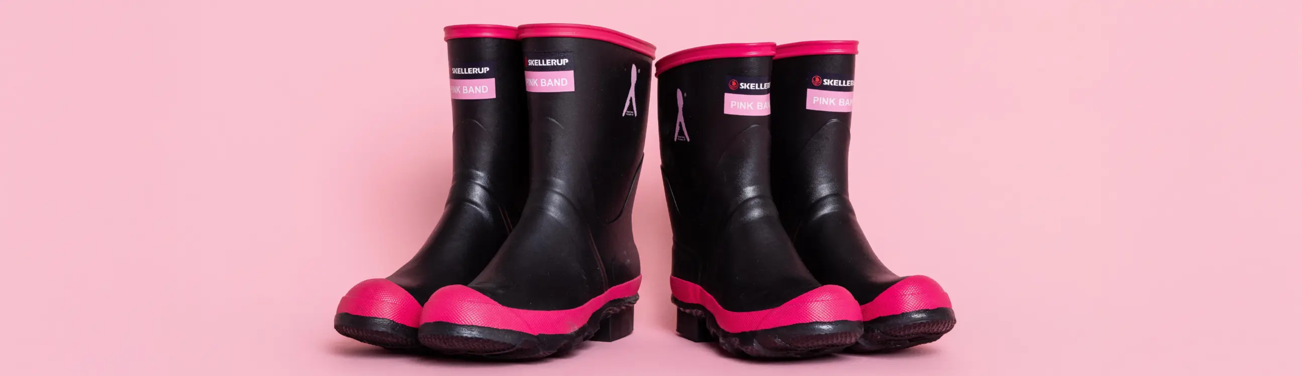 Two pairs of black rubber boots with pink soles and trim are placed side by side on a pink background. The boots have labels on the front.
