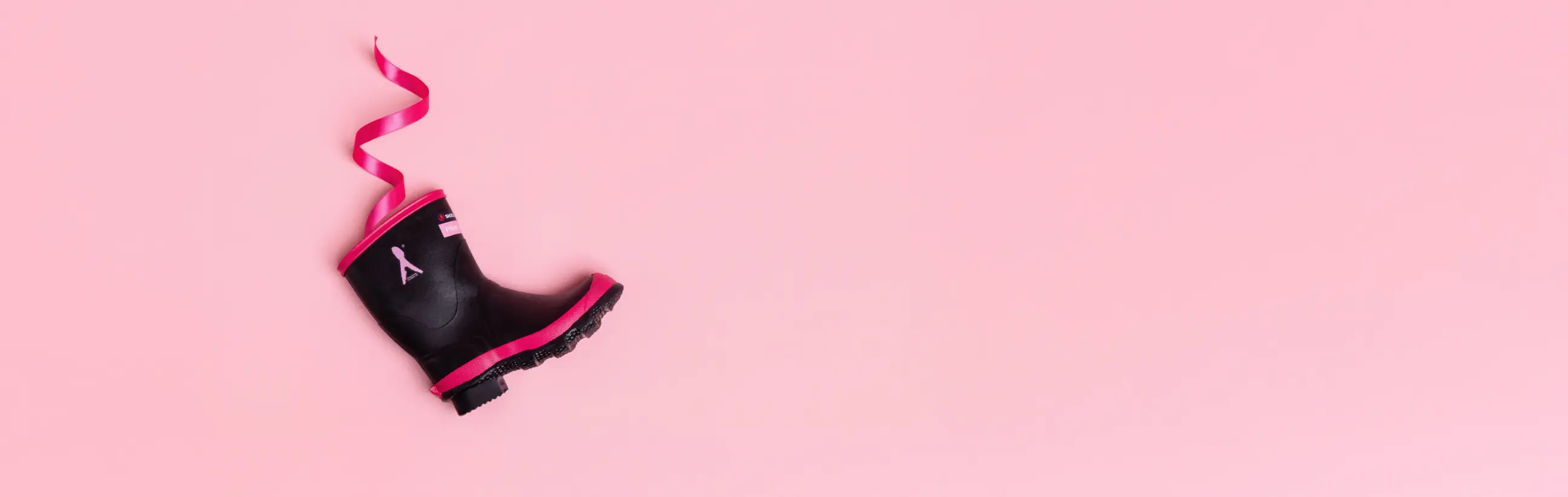 A single black rain boot with pink trim and a pink ribbon is positioned on a pastel pink background, with the ribbon curling upward from the boot.
