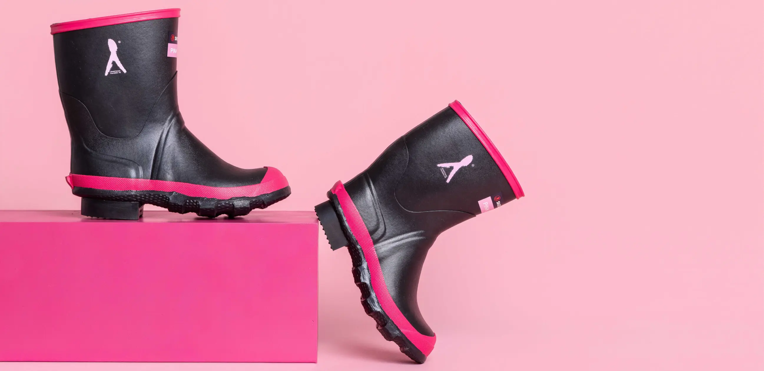 Two black rain boots with pink soles and trim are displayed against a pink background. One boot stands upright on a pink block, while the other leans at an angle against the block.