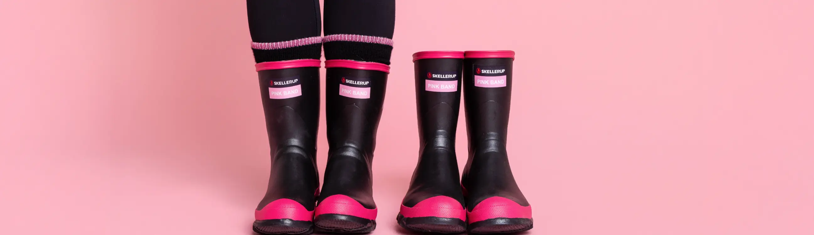 Two pairs of black and pink rain boots on a pink background, worn by two people standing side by side; only their lower legs and feet are visible.