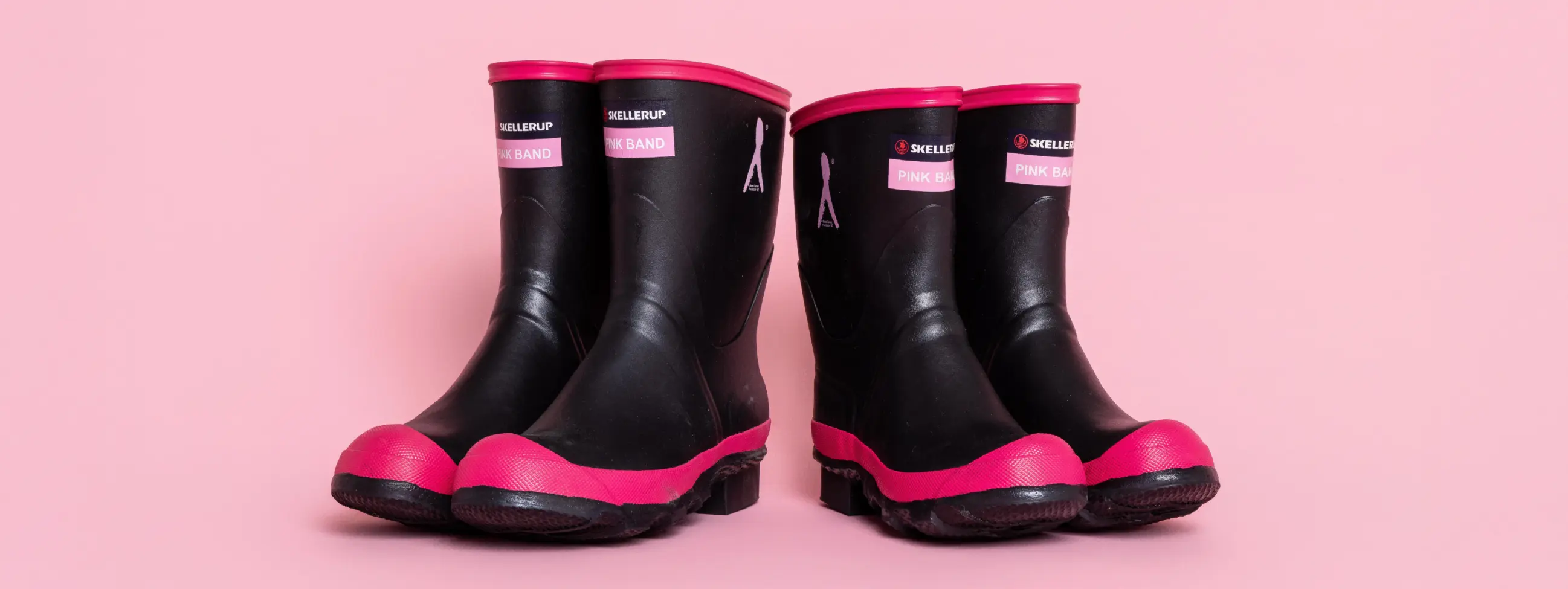 Two pairs of black rubber boots with pink soles and trim are placed side by side on a pink background. The boots have labels on the front.
