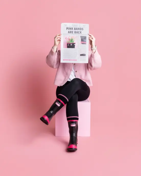 A person wearing black leggings, pink and black boots, and a pink cardigan sits on a pink cube against a pink background, holding up a newspaper that covers their face.