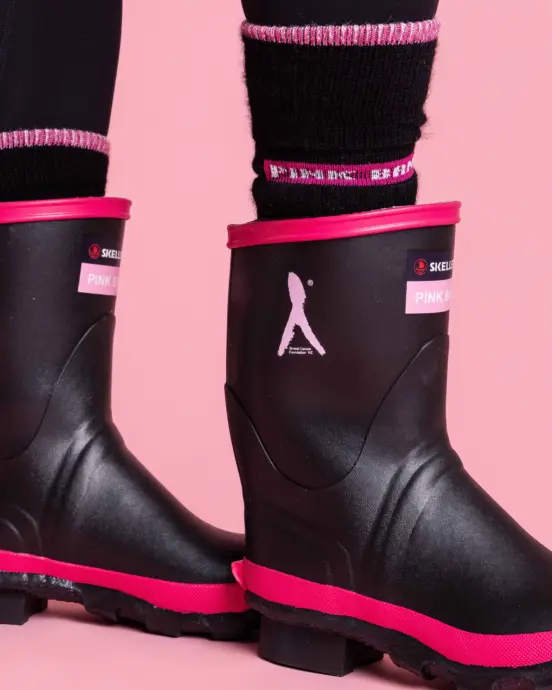 Two legs wearing black rain boots with pink trim and logo details, along with black socks featuring pink and white stripes at the top, standing on a pink background.