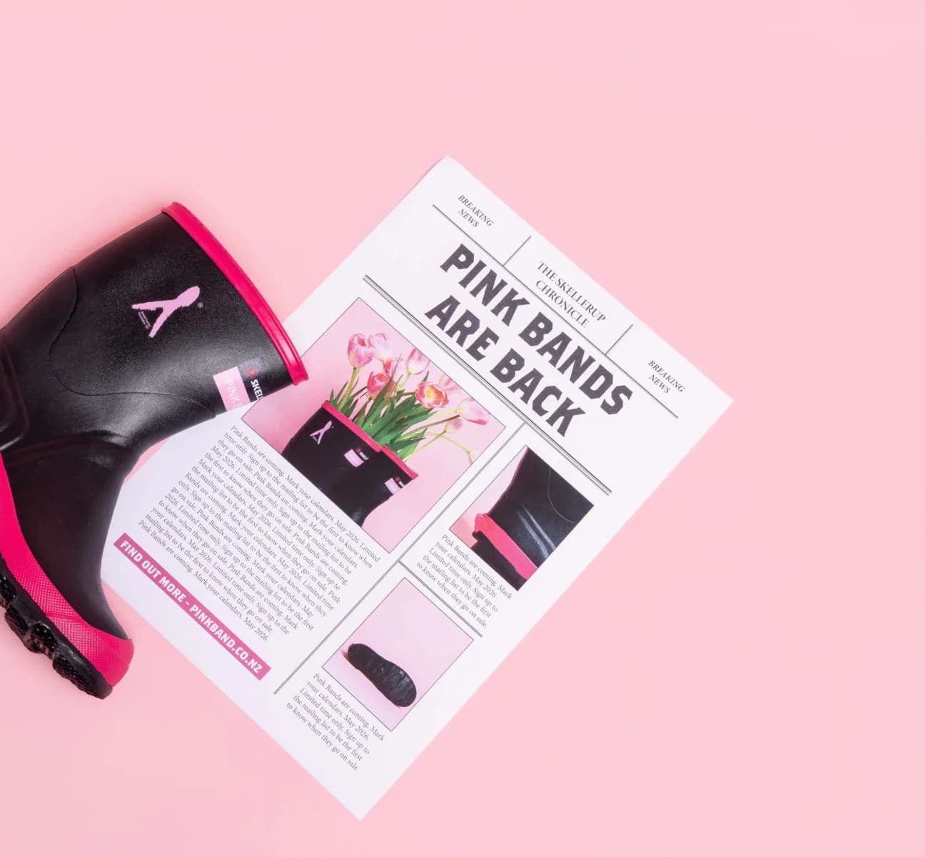 A black and pink rain boot with a pink ribbon symbol rests on a pink surface next to a newspaper titled PINK BANDS ARE BACK, featuring articles and images about the boots.
