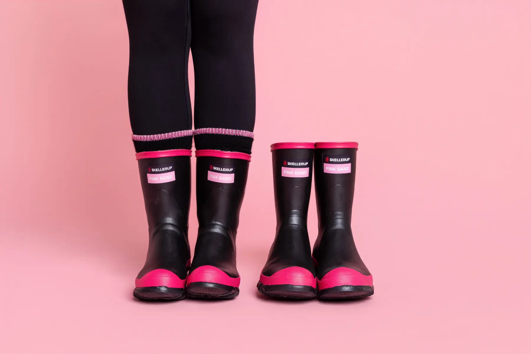 Two pairs of black and pink rain boots, one smaller and one larger, are worn by two people standing side by side against a pink background. Only their legs and feet are visible.