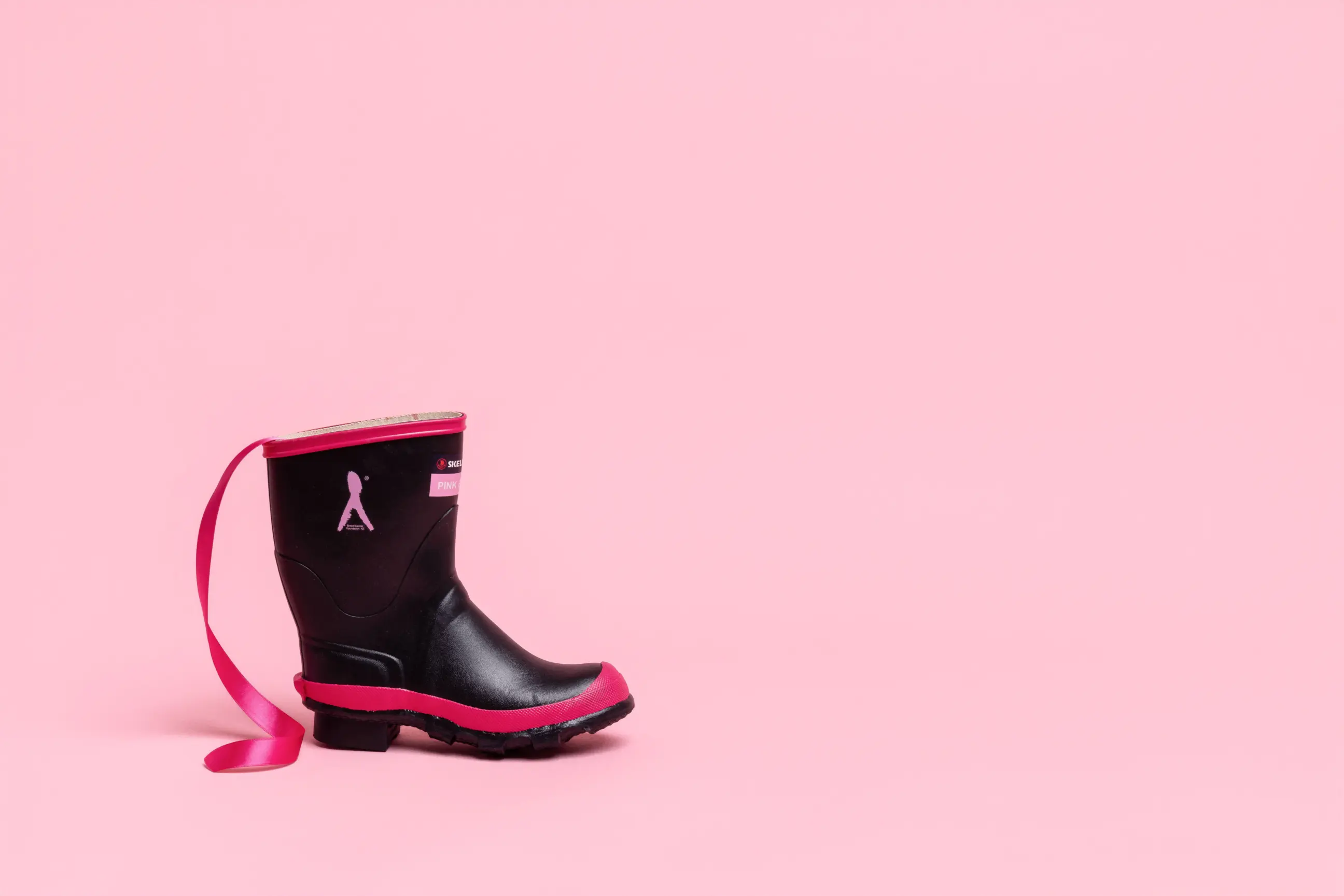 A single black rain boot with pink trim and a pink ribbon design sits on a light pink background. A loose pink ribbon extends from the boot, adding a pop of color. The scene is minimalistic with ample empty space.