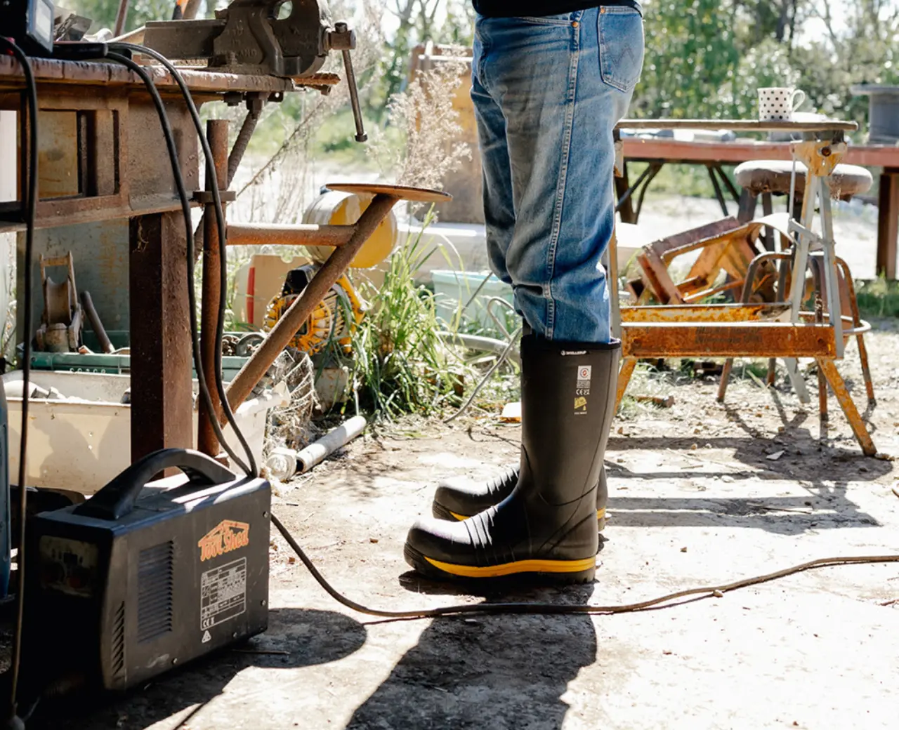 A person in jeans and black rubber boots works with tools at an outdoor workbench, surrounded by equipment and machinery, with sunlight casting shadows on the ground.