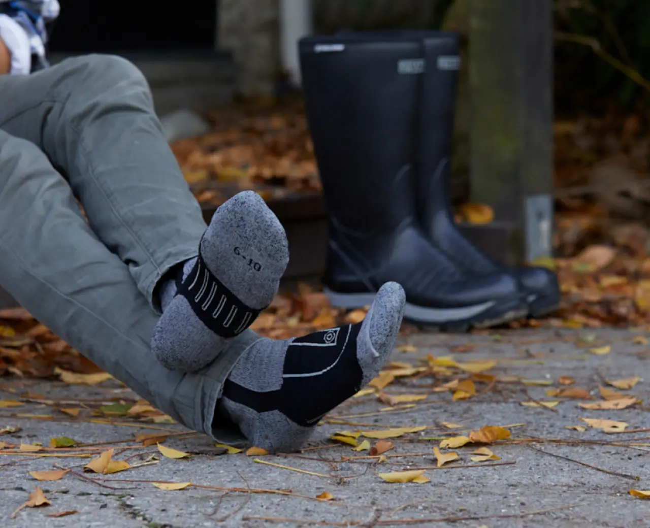 A person wearing grey and black socks sits on an outdoor surface covered with fallen leaves. Their boots are placed on the ground nearby. Only the persons legs and feet are visible.