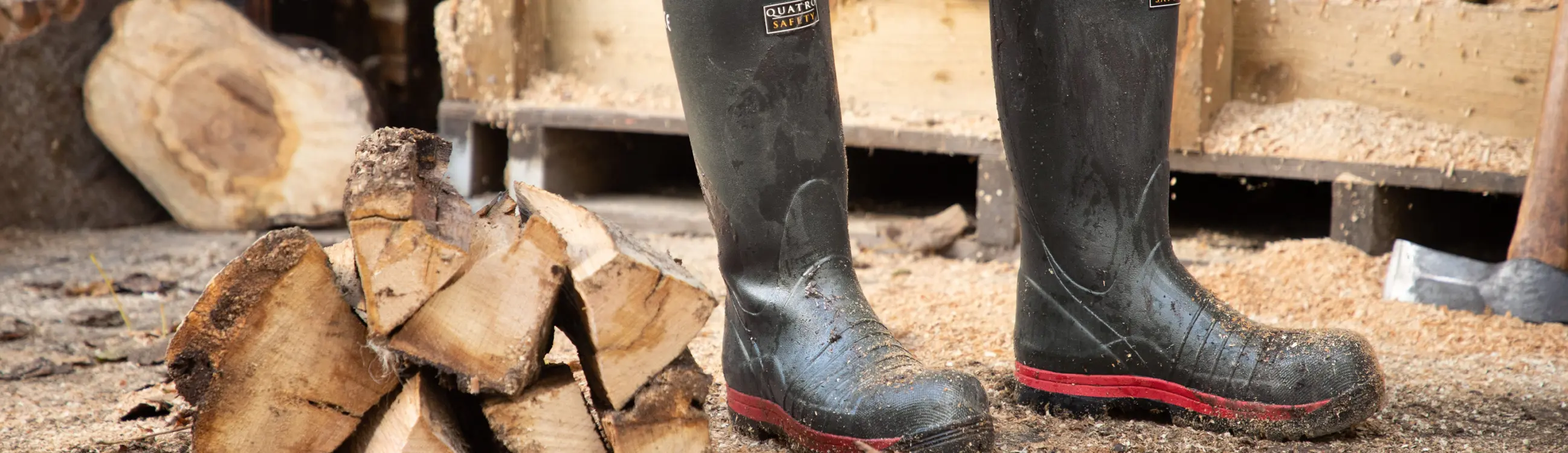 A pair of muddy black rubber boots stands on the ground near chopped firewood, with an axe and stacked logs in the background.