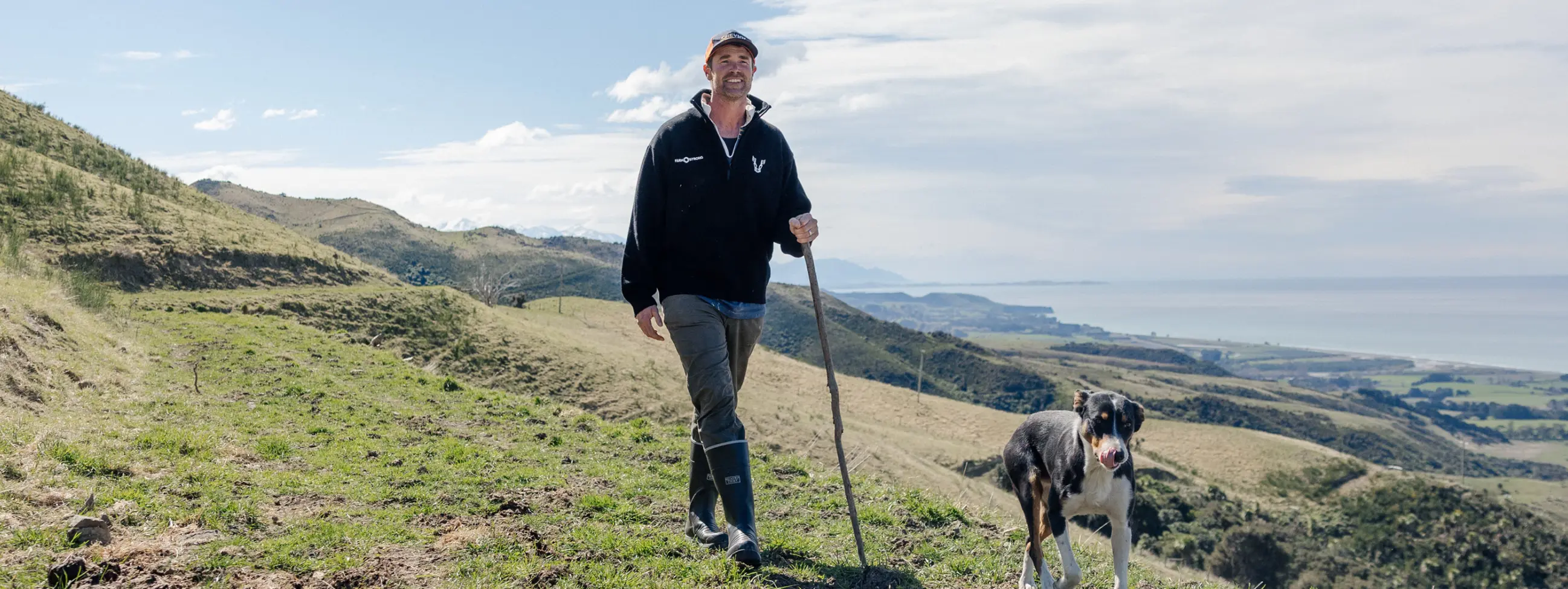 A person wearing a black hoodie and rubber boots stands on a grassy hill with a dog, holding a walking stick. Hills, coastline, and the ocean are visible in the background under a partly cloudy sky.