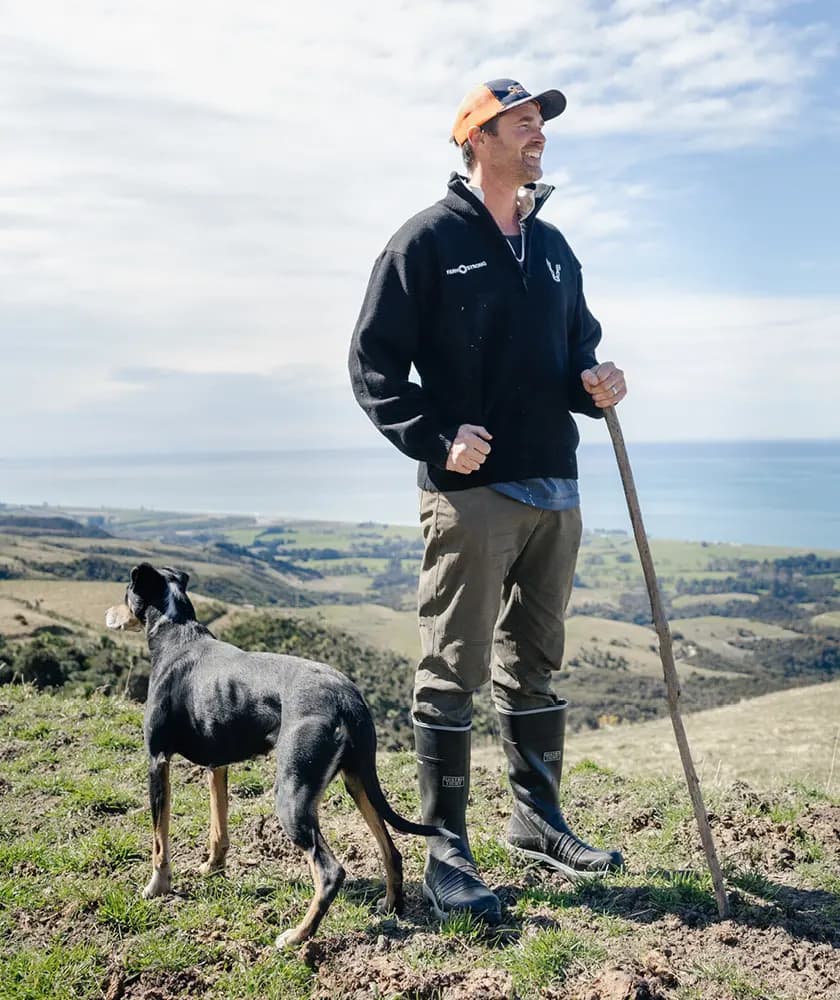 A man in outdoor clothing and rubber boots stands on a grassy hilltop with a dog and a wooden stick, overlooking a scenic landscape with rolling hills and the ocean in the distance under a partly cloudy sky.