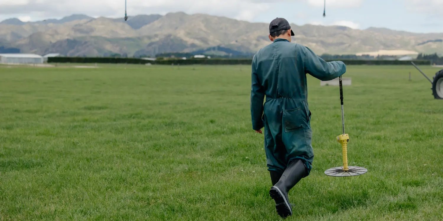 A person in green coveralls and boots walks across a grassy field, holding a circular measuring tool. The scene is outdoors with hills and clouds visible in the background.