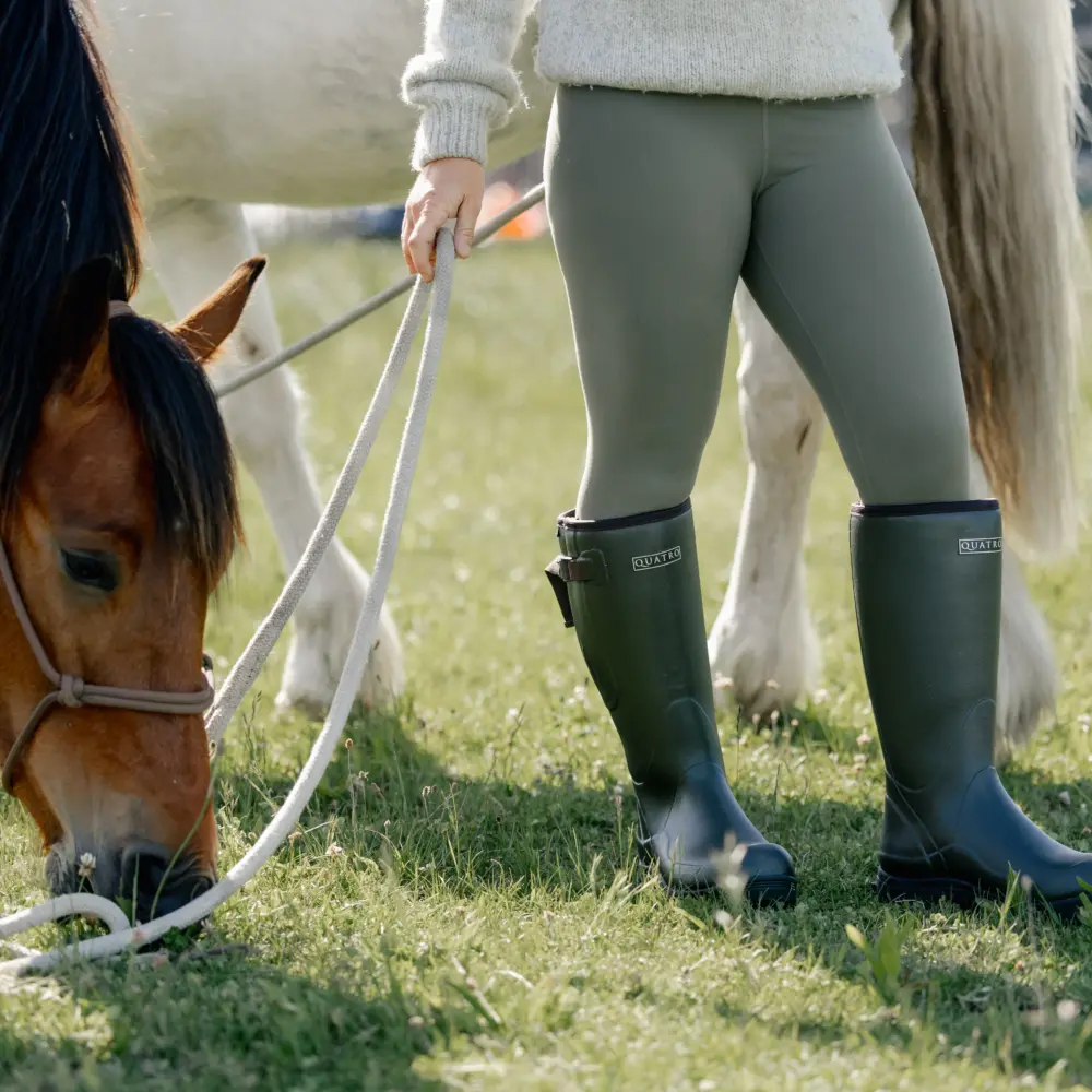 A person in green rubber boots and leggings holds a lead rope as a brown horse with a white blaze grazes on grass; a second white horse stands in the background.