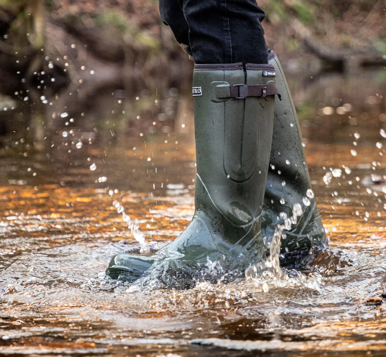 Person wearing tall green rubber boots splashes through shallow water, causing droplets to fly. The background is blurred, showing an outdoor natural setting.